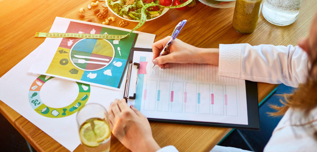   Woman dietitian in medical uniform with tape measure working on a diet plan sitting with different healthy food ingredients in the green office on background. Weight loss and right nutrition concept
